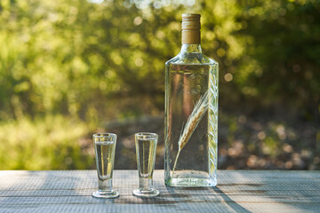 Bottle of crystal clear, famous and delicious Polish vodka with macerated corn ear inside and two old vintage glasses , served outdoor in summer sunny day. Popular drink in eastern european countries. - obrazy, fototapety, plakaty