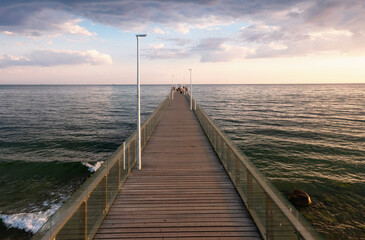 Fototapeta na wymiar Perspective view of a long sea pier. There is a boardwalk and glass railings. Morning seascape. Human figures are visible in the distance. Background.
