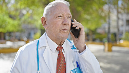 Senior grey-haired man doctor standing with serious expression talking on smartphone at park