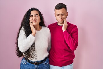 Naklejka premium Young hispanic couple standing over pink background touching mouth with hand with painful expression because of toothache or dental illness on teeth. dentist concept.