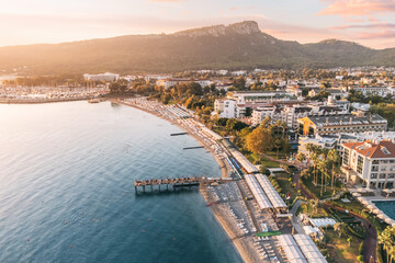 Naklejka premium Indulge in a captivating visual journey with mesmerizing aerial shot, revealing a panoramic vista of Kemer's hotels harmoniously nestled between a picturesque sea beach and awe-inspiring mountains.