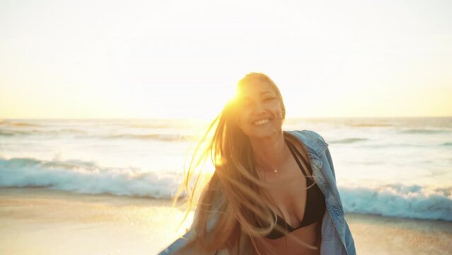 Smiling woman spinning on ocean beach during sunset. Blonde hair model in denim shirt and bikini enjoying freedom on summer vacation and sending blow kiss camera. Concept joy and serenity