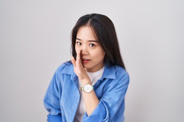 Young chinese woman standing over white background hand on mouth telling secret rumor, whispering malicious talk conversation