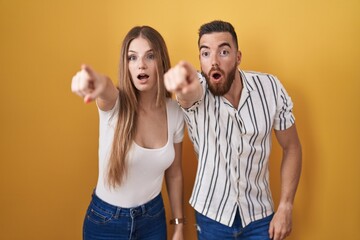 Young couple standing over yellow background pointing with finger surprised ahead, open mouth...