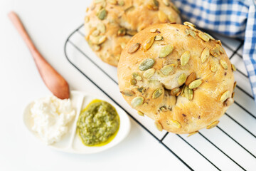 A freshly baked bagel with sunflower seeds lies on a metal stand along with a plate of cheese and pesto sauce and a wooden knife.