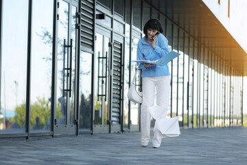 Fototapeta premium Multitasking busy young business woman communicates by smartphone during walks reading and dropping important finance documents by the office buildings outdoors. Stress resistance and business concept