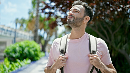 Young hispanic man tourist wearing backpack looking around smiling at park