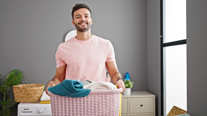 Young hispanic man smiling confident holding basket with clothes at laundry room