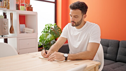 Young hispanic man writing on notebook sitting on table at dinning room
