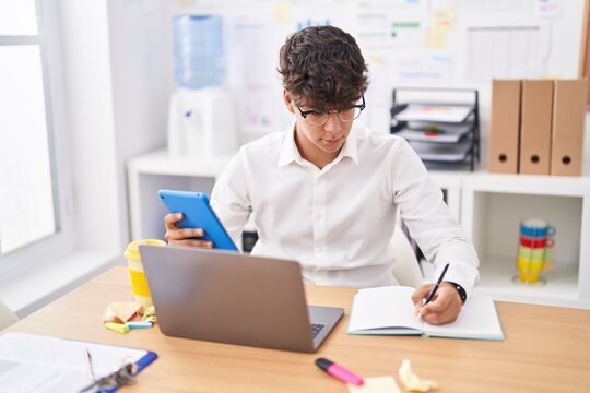 Young hispanic teenager business worker using touchpad writing on notebook at office