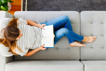 Top view of young brunette woman relaxing on sofa and reading novel book at home