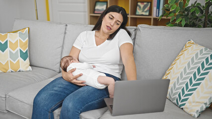 Mother and baby daughter sitting on sofa holding baby using smartphone and laptop at home