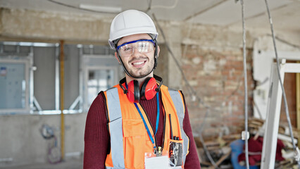 Young hispanic man builder smiling confident standing at construction site