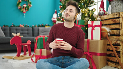 Obraz na płótnie Canvas Young hispanic man listening to music drinking coffee celebrating christmas at home