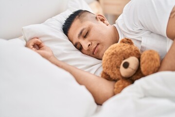 Young latin man hugging teddy bear lying on bed sleeping at bedroom