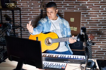 Hispanic young man playing classic guitar at music studio showing middle finger, impolite and rude fuck off expression