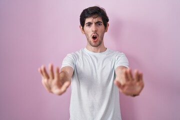 Naklejka premium Young hispanic man standing over pink background doing stop gesture with hands palms, angry and frustration expression