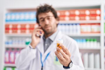 Young hispanic man pharmacist holding pills bottle talking on smartphone at pharmacy