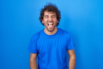 Fototapeta na wymiar Hispanic young man standing over blue background sticking tongue out happy with funny expression. emotion concept.