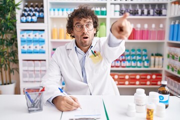 Hispanic young man working at pharmacy drugstore pointing with finger surprised ahead, open mouth amazed expression, something on the front