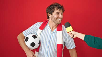 Young hispanic man supporting soccer team having interview over isolated red background