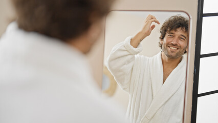 Young hispanic man wearing bathrobe combing hair with hands looking on mirror at home