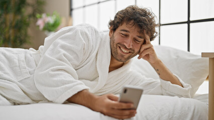 Young hispanic man wearing bathrobe using smartphone smiling at bedroom - obrazy, fototapety, plakaty
