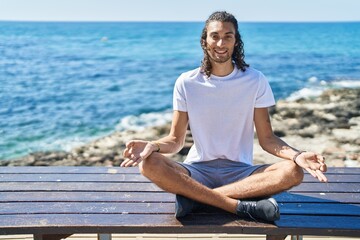 Young hispanic man doing yoga exercise sitting on bench at seaside