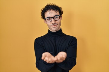 Hispanic man standing over yellow background smiling with hands palms together receiving or giving gesture. hold and protection