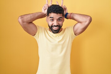 Hispanic man with beard standing over yellow background posing funny and crazy with fingers on head as bunny ears, smiling cheerful
