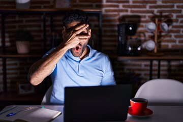 Hispanic man with beard using laptop at night peeking in shock covering face and eyes with hand, looking through fingers with embarrassed expression.