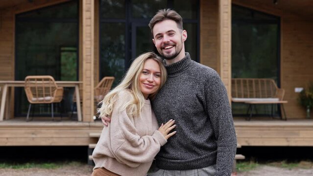 Portrait of happy couple hugging looking at camera standing on yard of wooden summer house. Front view medium shot Caucasian handsome man and beautiful woman posing on weekend enjoying leisure