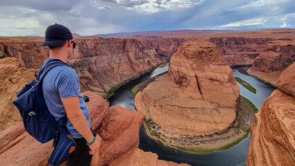 Tourist man standing at the cliff edge with panoramic aerial view of Horseshoe bend on the Colorado...