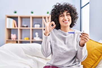 Young brunette woman with curly hair holding pregnancy test result doing ok sign with fingers, smiling friendly gesturing excellent symbol