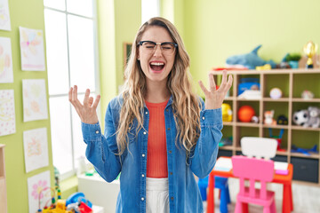 Young caucasian woman working as teacher at kindergarten crazy and mad shouting and yelling with...