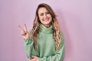 Young caucasian woman standing over pink background smiling with happy face winking at the camera doing victory sign. number two.