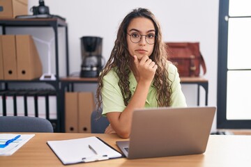 Young hispanic woman working at the office wearing glasses looking confident at the camera with smile with crossed arms and hand raised on chin. thinking positive.