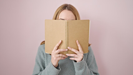 Young blonde woman covering face with book over isolated pink background