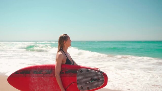 Surfing on summer vacation. Female tourist in black bikini holding red surfboard and walking into ocean. Woman practicing her surfing skills. Healthy active lifestyle