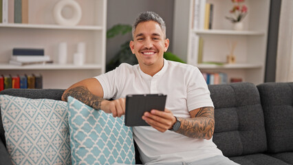 Young hispanic man using touchpad sitting on sofa at home