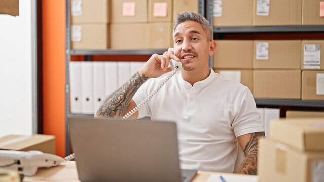 Young hispanic man ecommerce business worker using laptop talking on telephone smiling at office