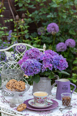 Tischdekoration mit Porzellan-Kaffeetasse, Cookies und lila Hortensie im Krug