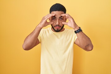 Fototapeta na wymiar Young hispanic man standing over yellow background trying to open eyes with fingers, sleepy and tired for morning fatigue