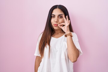 Young arab woman standing over pink background mouth and lips shut as zip with fingers. secret and...