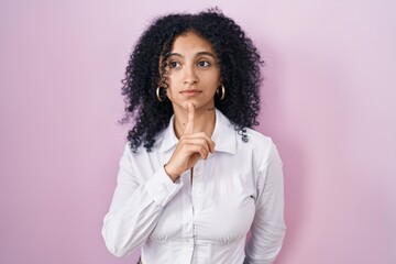 Fototapeta na wymiar Hispanic woman with curly hair standing over pink background thinking concentrated about doubt with finger on chin and looking up wondering