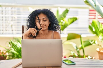 Young hispanic woman using laptop sitting on table with serious expression at home terrace