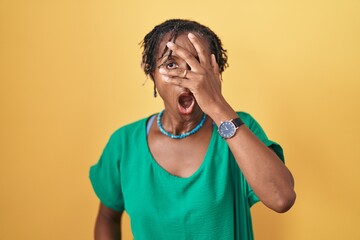 African woman with dreadlocks standing over yellow background peeking in shock covering face and...