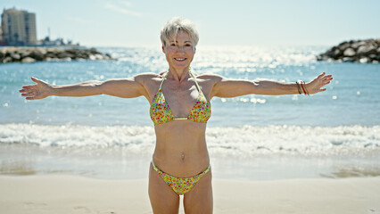 Middle age blonde woman tourist wearing bikini smiling confident with arms open at the beach