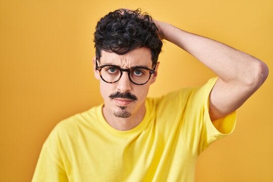 Hispanic man wearing glasses standing over yellow background confuse and wonder about question. uncertain with doubt, thinking with hand on head. pensive concept.