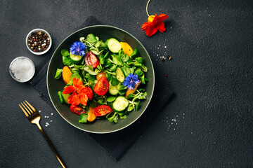 Green salad with fresh cucumbers, tomato, peach and nasturtium flowers on black background. Top view